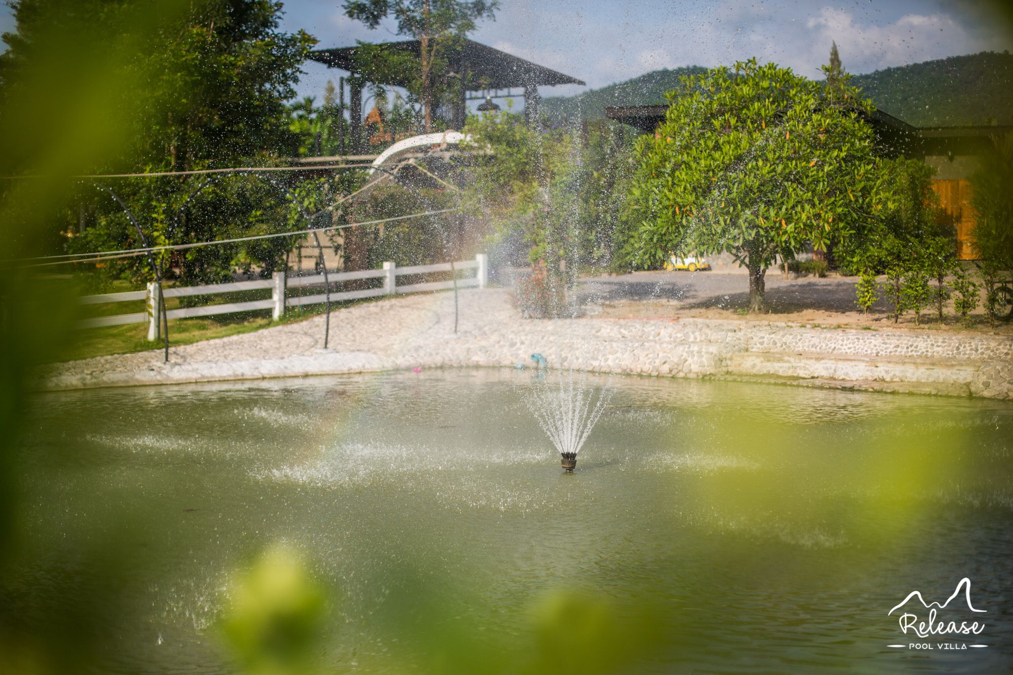 Una fuente en un parque con una cerca blanca y árboles al fondo.