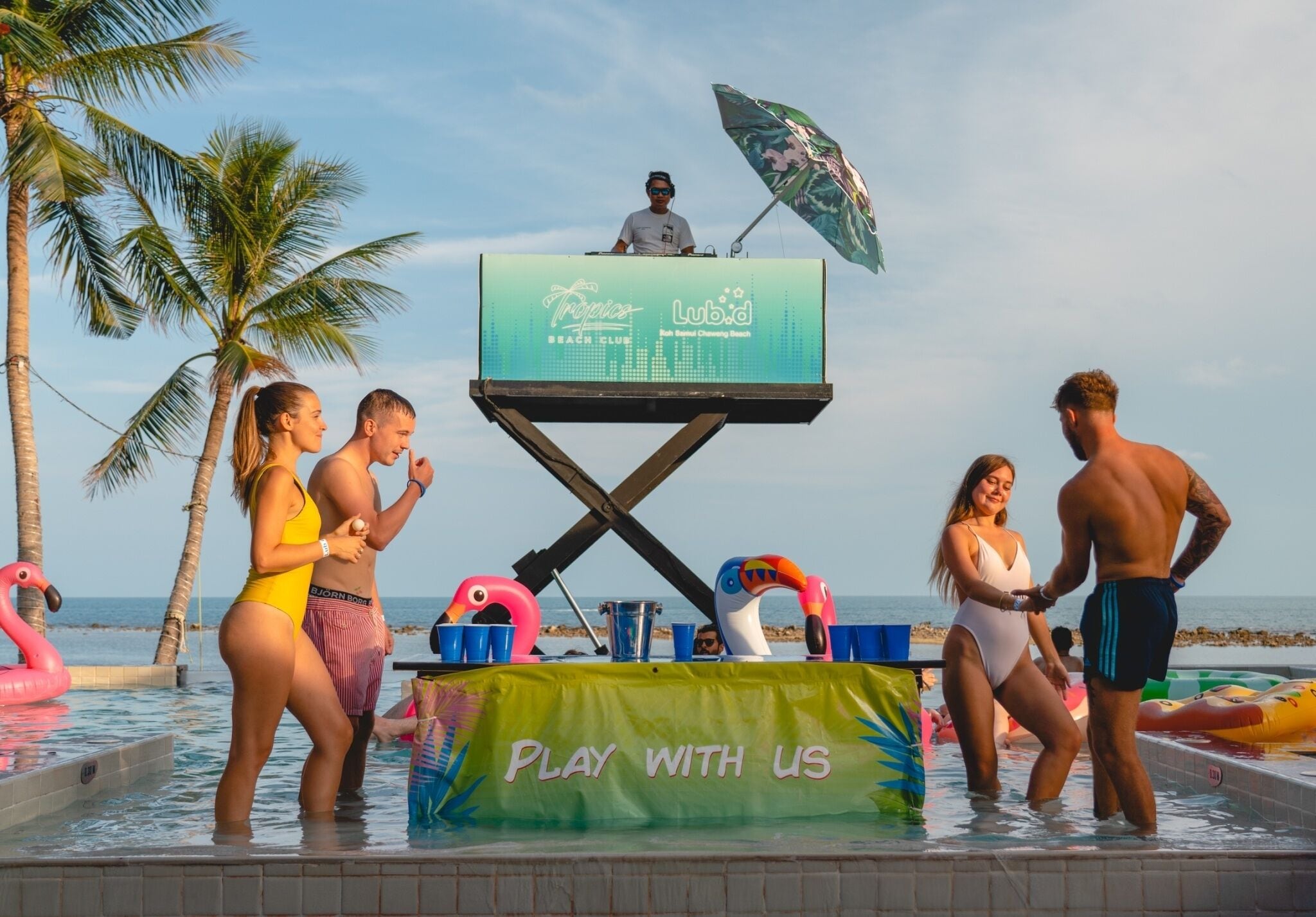 A poolside area with a DJ booth, inflatable flamingos, and a banner that reads "Play with us".