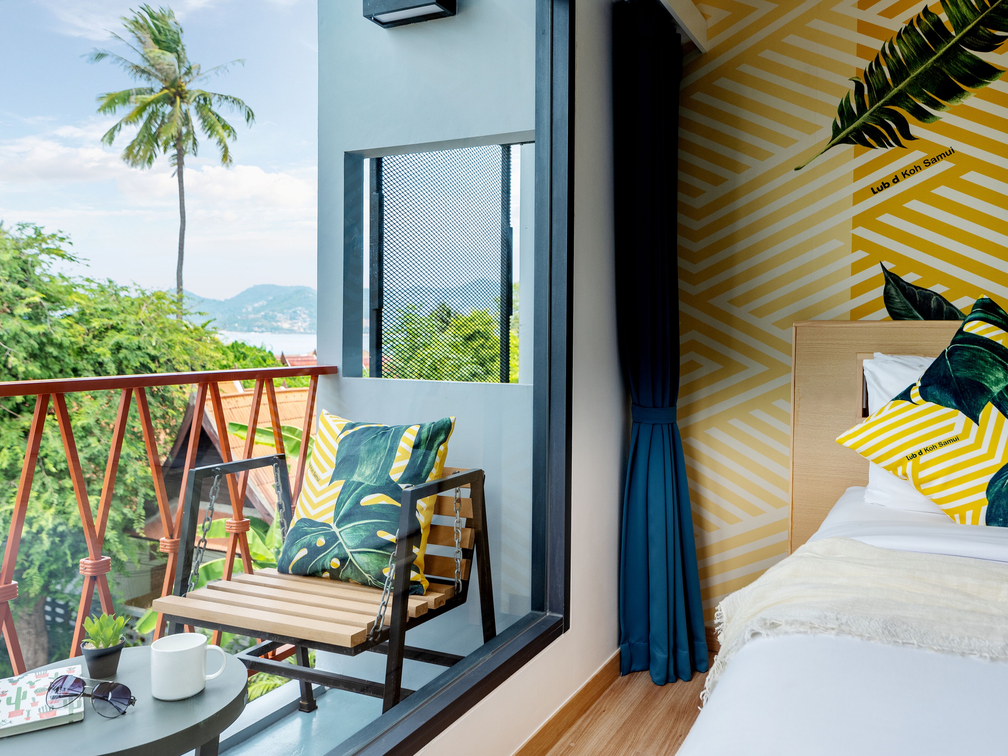 A balcony with a wooden chair, a small table with a cup and sunglasses, and a view of palm trees and mountains.