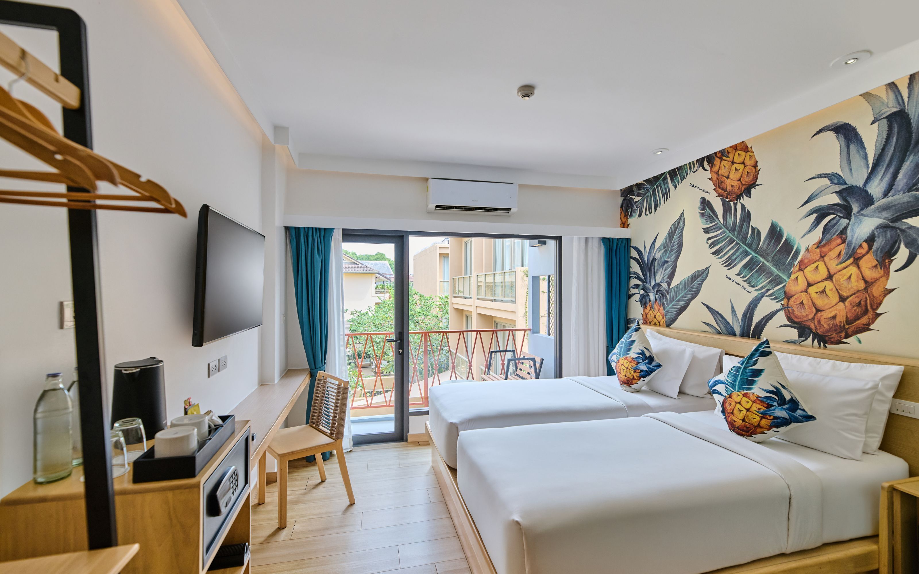 A hotel room with two beds, a desk, a TV, and a balcony with a view of the city.