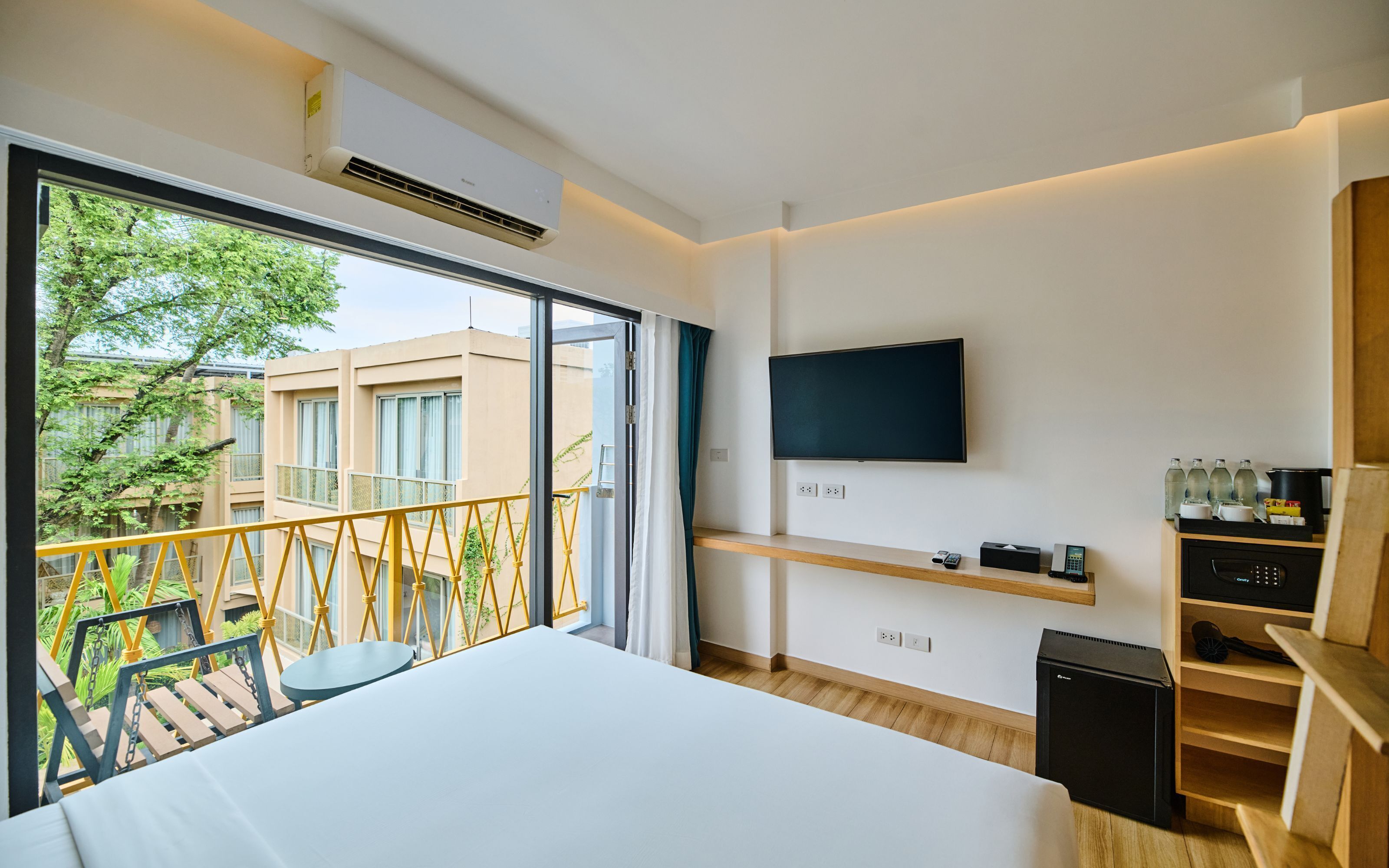 A modern hotel room with a large bed, a TV mounted on the wall, and a balcony with a yellow railing.
