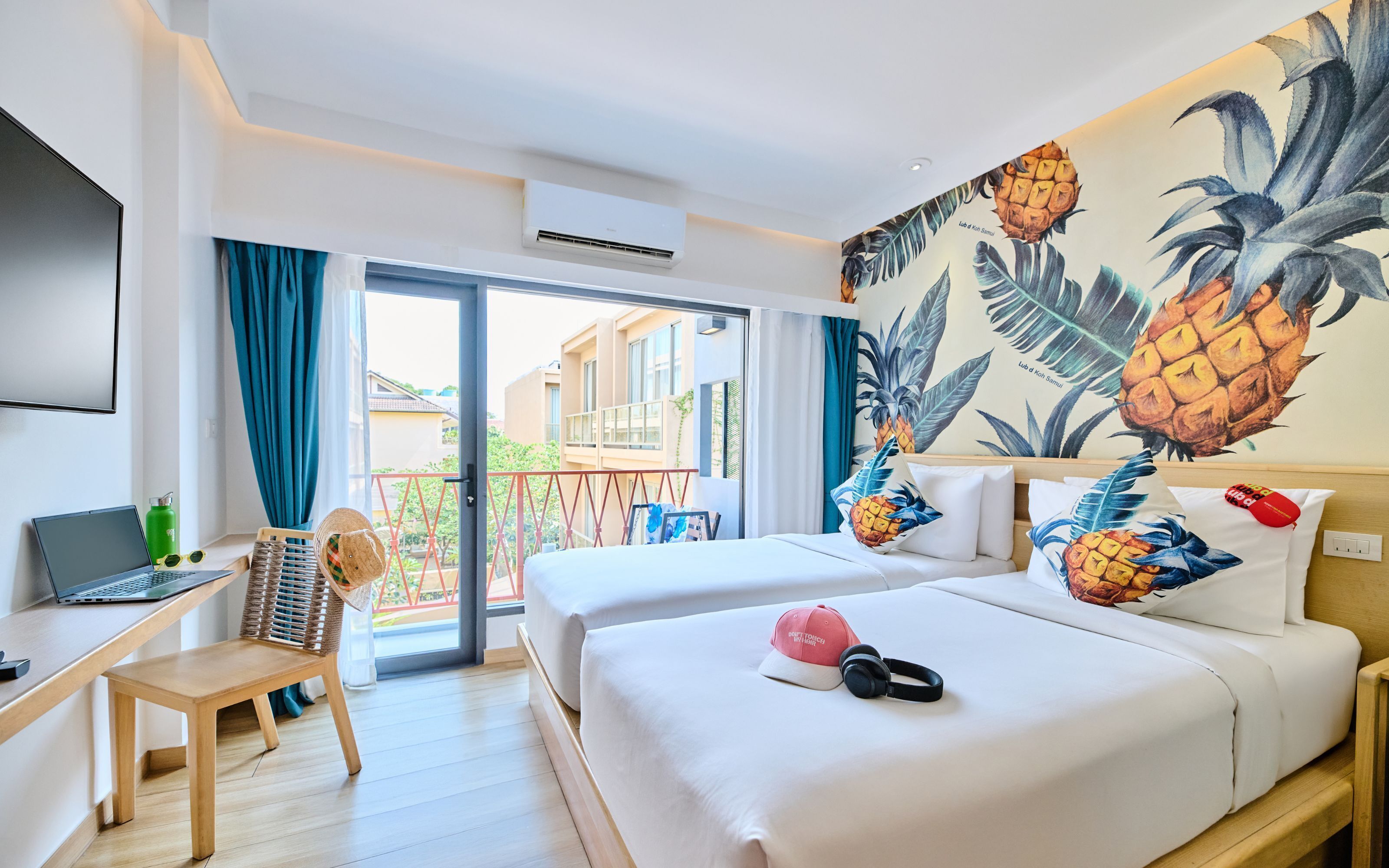 Une chambre d’hôtel avec deux lits, un bureau avec un ordinateur portable, une chaise et une grande fresque murale sur le thème des pays tropicaux.