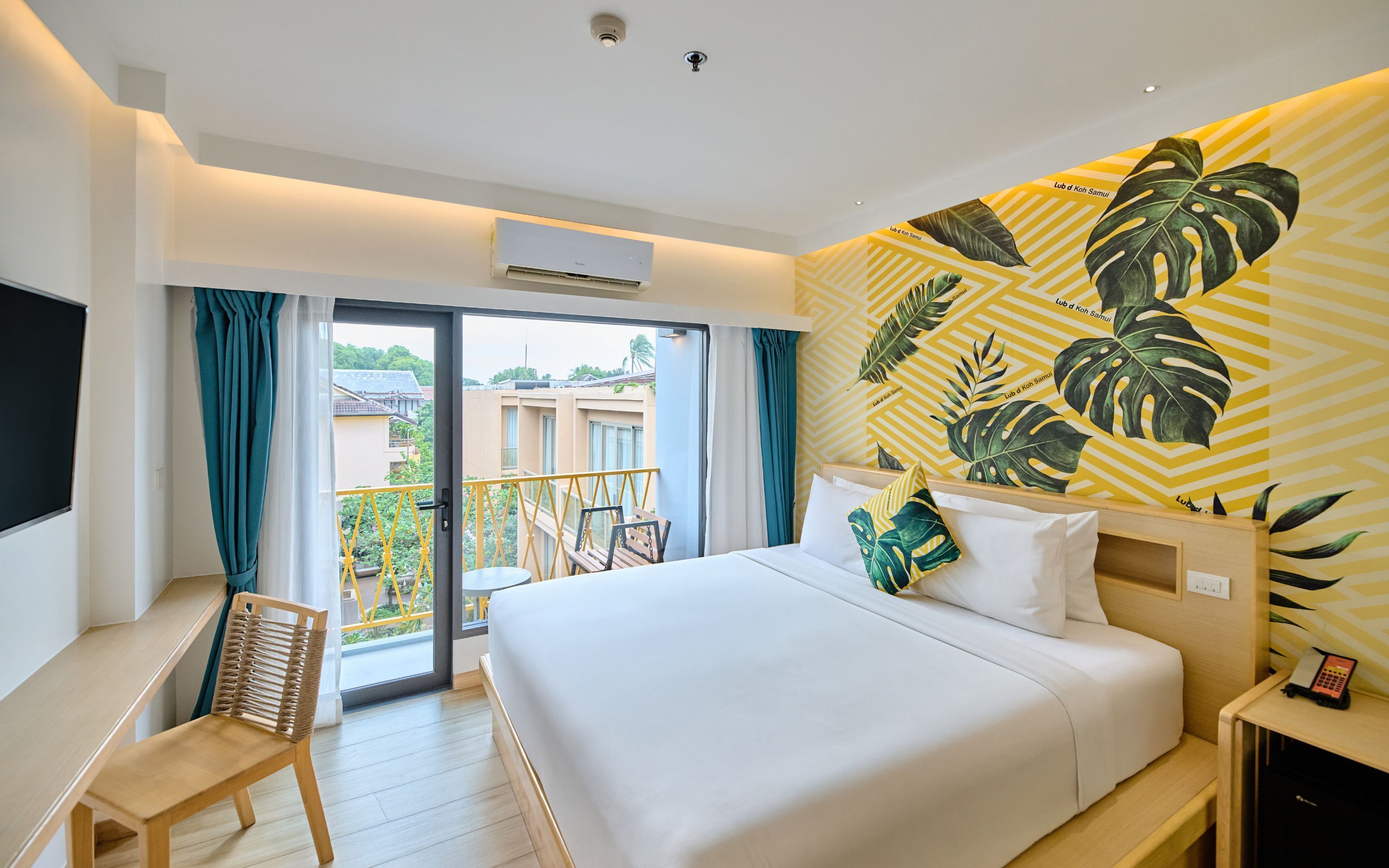 Ein Hotelzimmer mit Bett, Schreibtisch, Stuhl und einem Balkon mit gelben Geländern sowie einem großen Fenster.