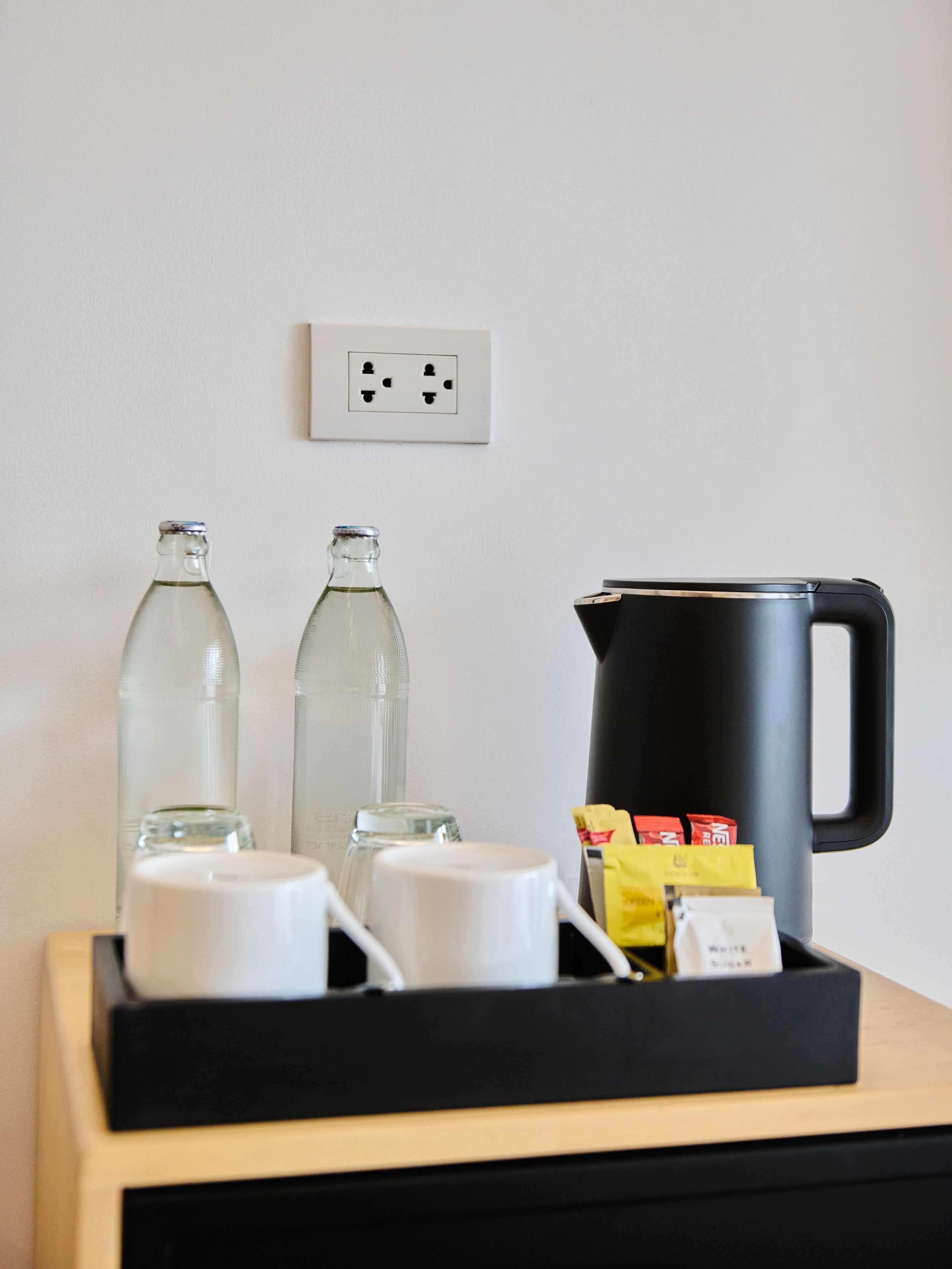 A kitchenette area with a black kettle, two glass bottles, and a white mug on a wooden counter.