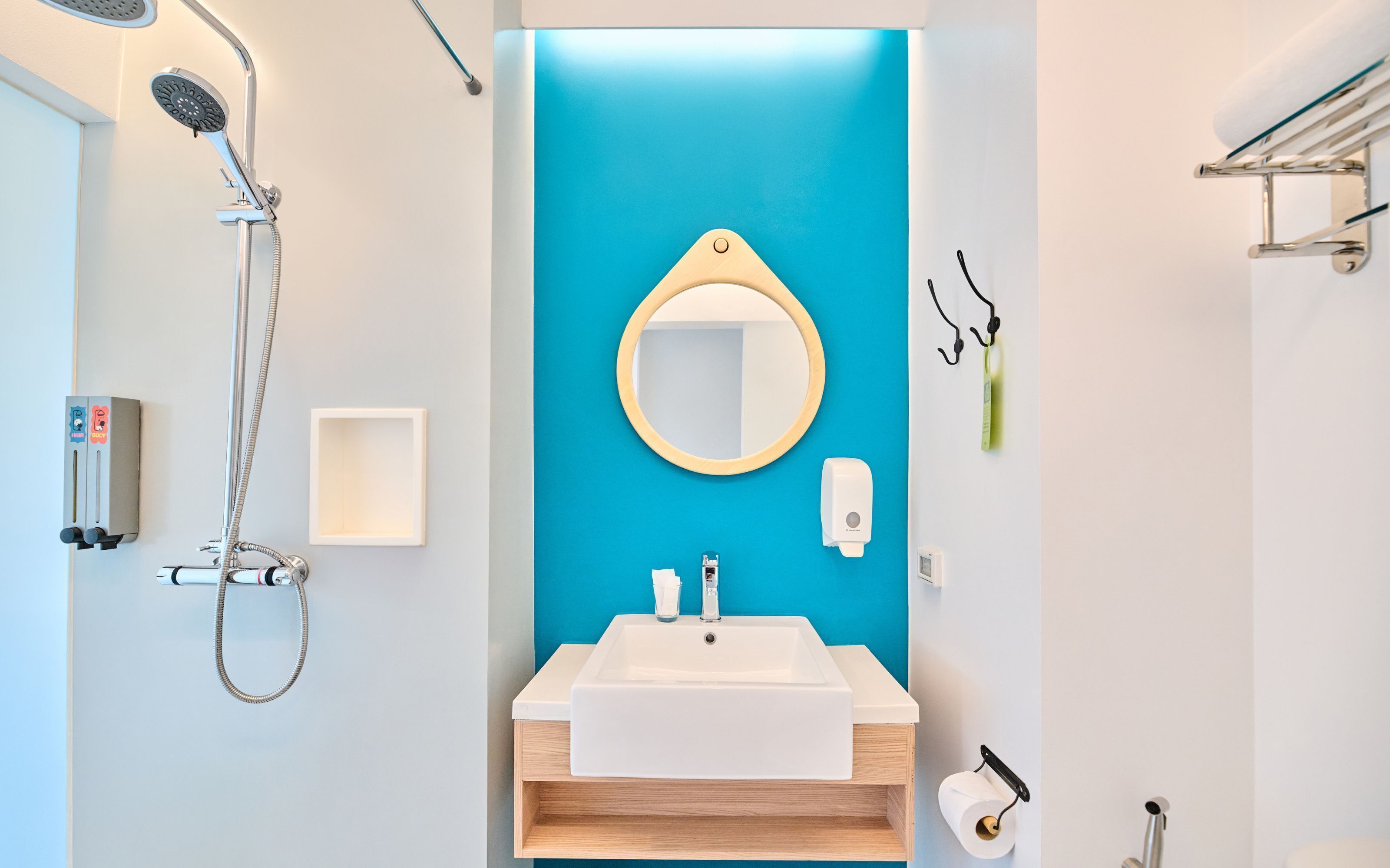 A modern bathroom with a blue accent wall, a white sink, a mirror, a handheld showerhead, a soap dispenser, and a toilet paper holder.
