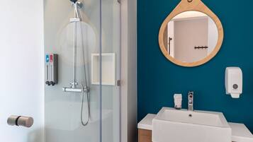 A modern bathroom with a glass-enclosed shower, a white sink with a wooden vanity, and a round mirror on a teal wall.