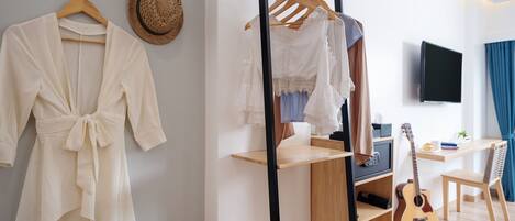 A room with a wooden floor, a bed with a headboard, a hanging clothes rack, a guitar, and a television on the wall.