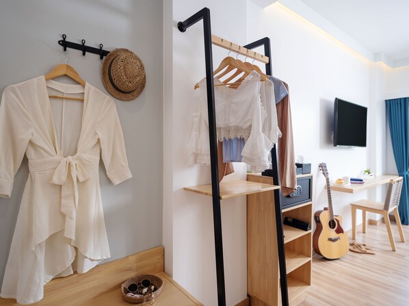 A room with a wooden floor, a bed with a headboard, a hanging clothes rack, a guitar, and a television on the wall.