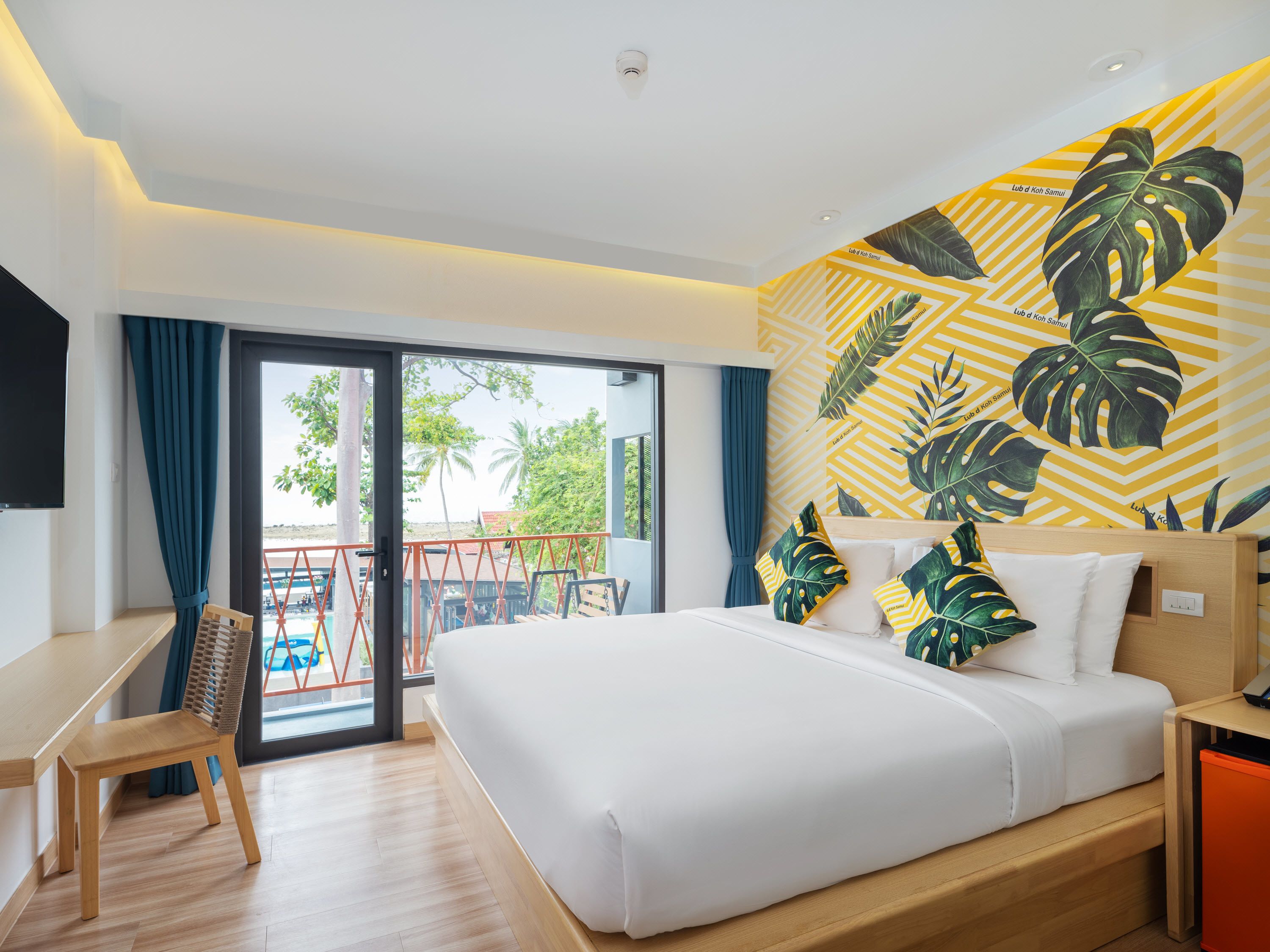 Habitación de hotel con una cama, un escritorio y una silla. Cuenta con un balcón que ofrece vistas a palmeras y una piscina.