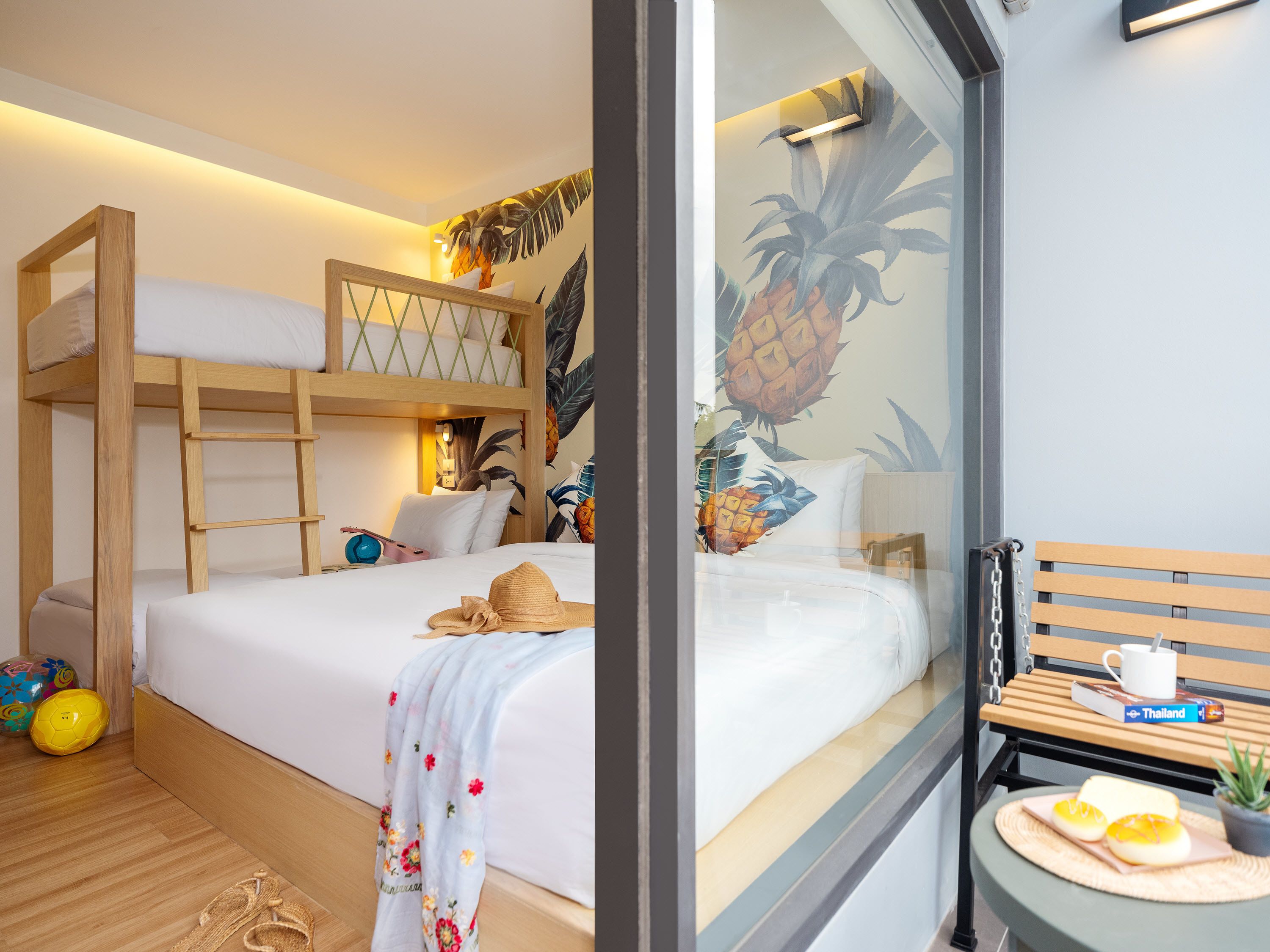 Um quarto com uma cama de solteiro, uma pequena mesa com um livro e uma xícara, e uma parede com um mural de abacaxi.