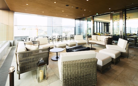 A modern lounge area with wicker furniture, a round table, and a view of the city through large windows.