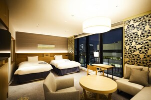 Ein Hotelzimmer mit zwei Betten, einem Sofa, einem Tisch und einem großen Fenster mit Blick auf die Stadt.