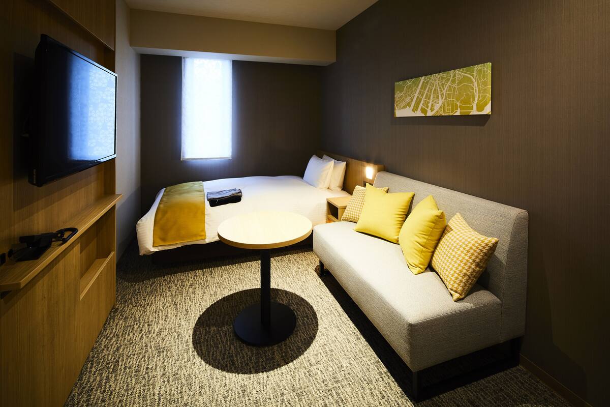 Una habitación de hotel moderna con sofá, una mesita redonda, televisión de pantalla plana y ventana que proporciona luz natural.