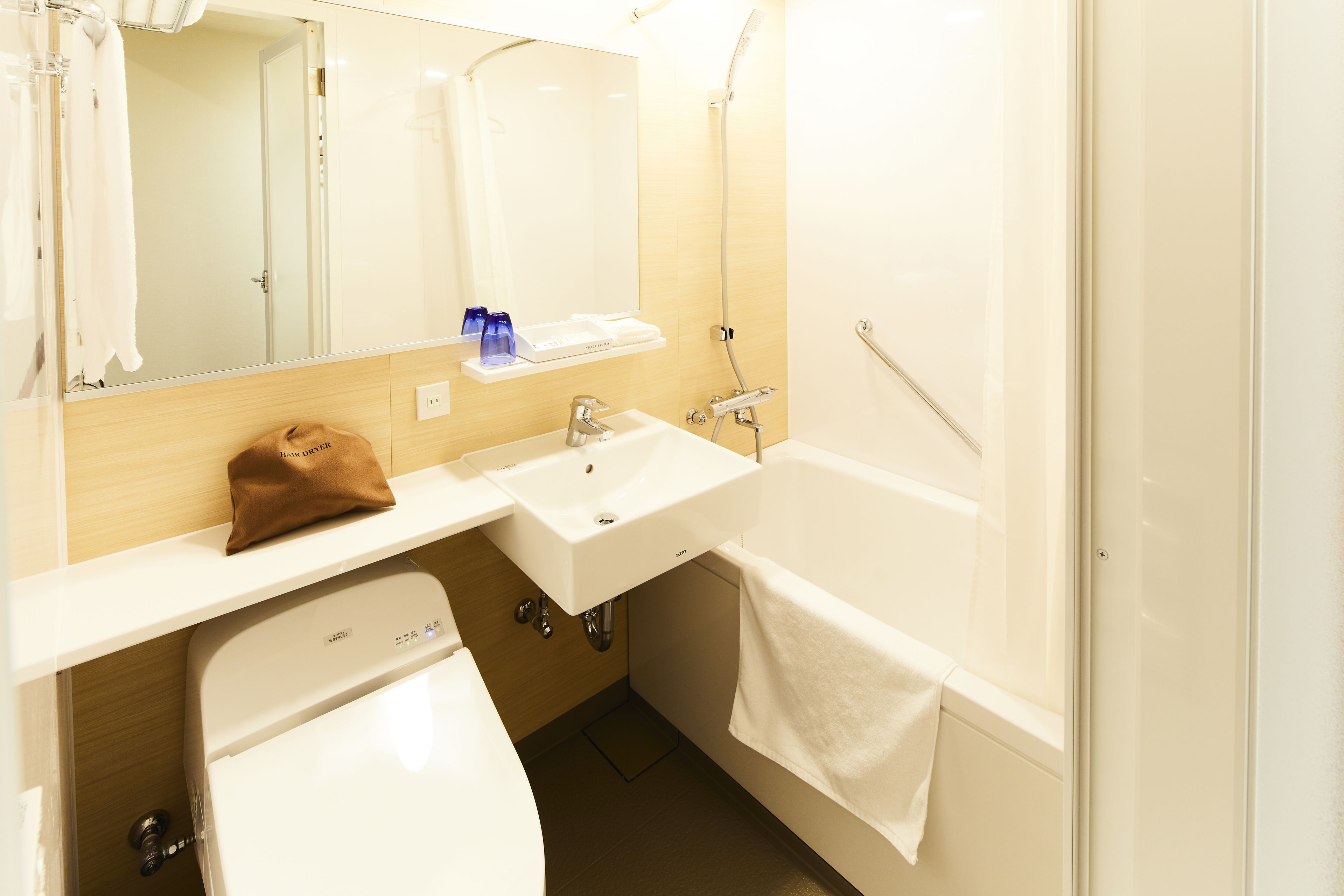 A bathroom with a white sink, mirror, and bathtub. There is a towel hanging on the wall and a brown bag on the counter.
