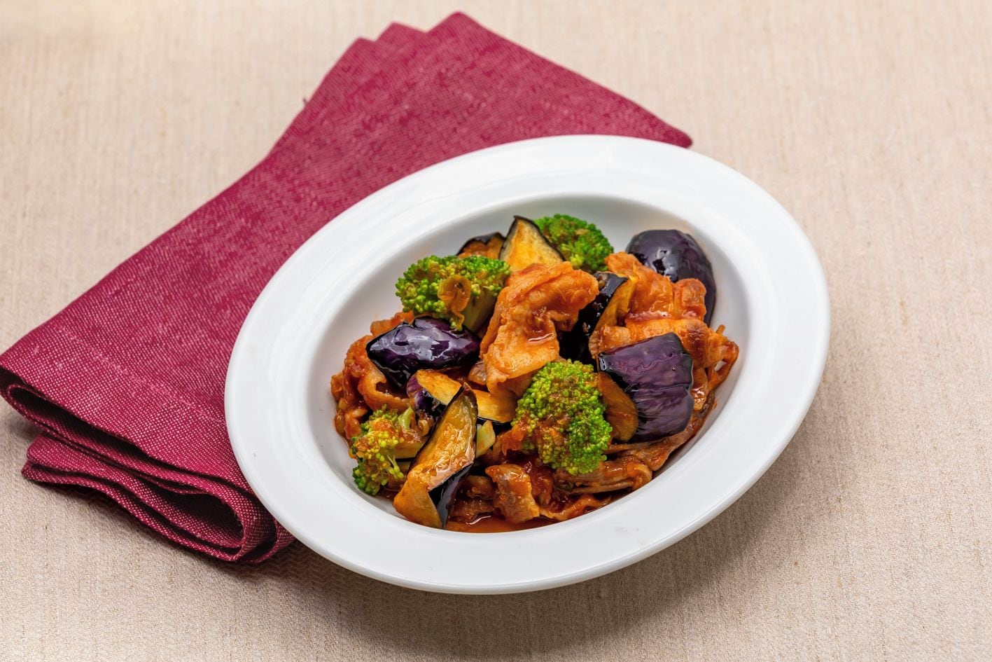 A bowl of stir-fried vegetables with broccoli and eggplant on a white plate, placed on a beige surface with a red cloth napkin beside it.