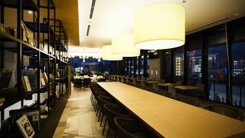 A modern bar area with a long wooden counter, multiple seating options, and a bookshelf with various items.