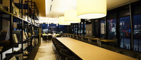A modern bar area with a long wooden counter, multiple seating options, and a bookshelf with various items.