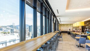 Zona de bar moderna con ventanas altas, una barra de madera larga y varios taburetes.