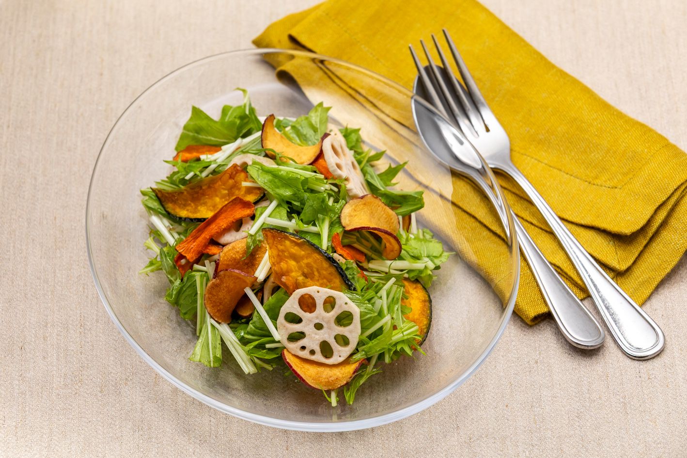 A clear glass bowl containing a salad with various greens and slices of vegetables, accompanied by a yellow napkin and silverware.