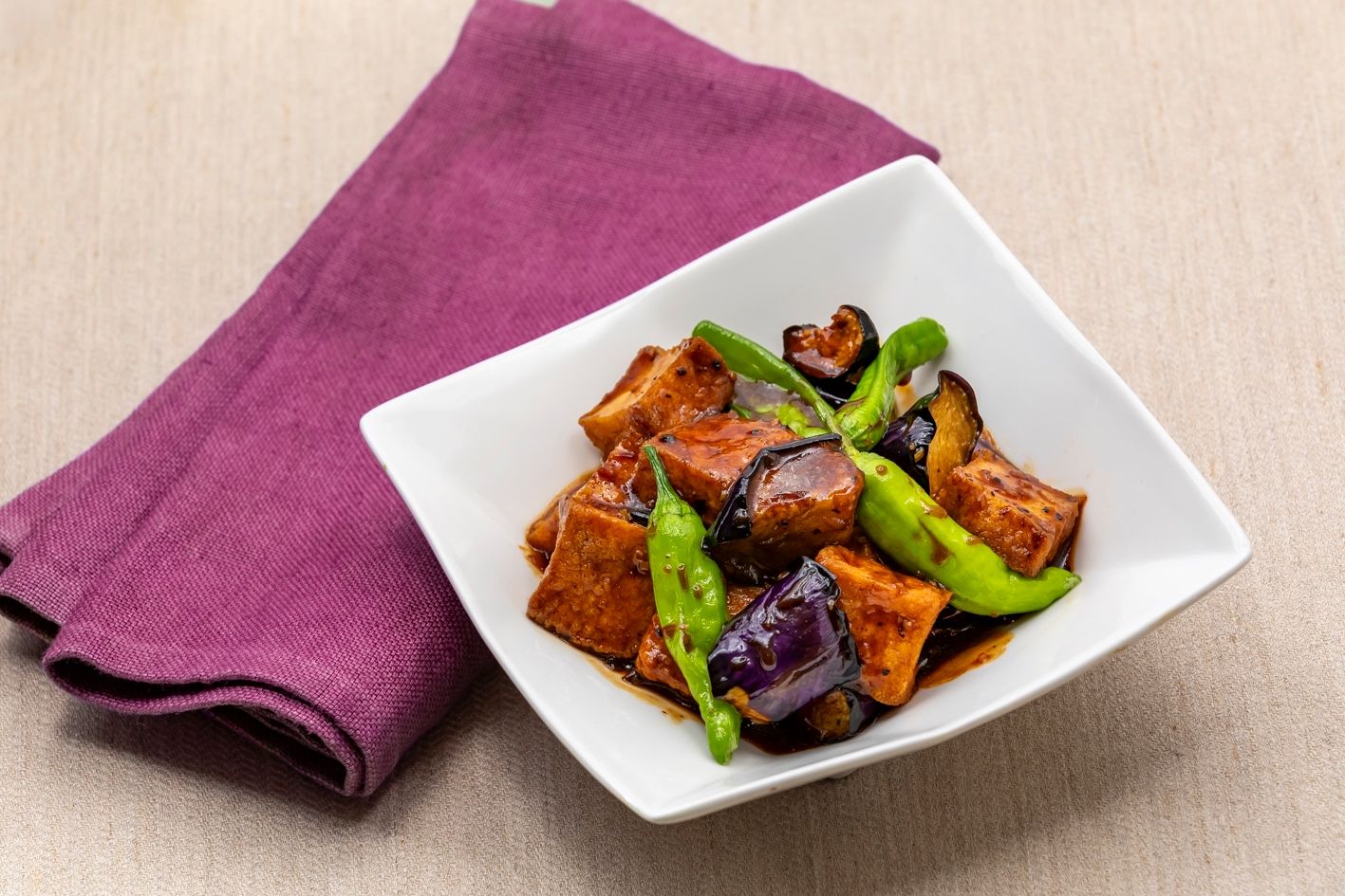 Un plato blanco cuadrado con un guiso, posiblemente tofu, con verduras verdes y moradas, sobre una superficie color beige con un paño púrpura.