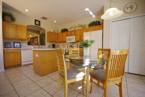 A kitchen with wooden cabinets, a white refrigerator, a microwave, a glass dining table with chairs, and a clock on the wall. - Ly53780 - Emerald Island - 7 Bed 4.5 Baths Villa (Kissimmee)