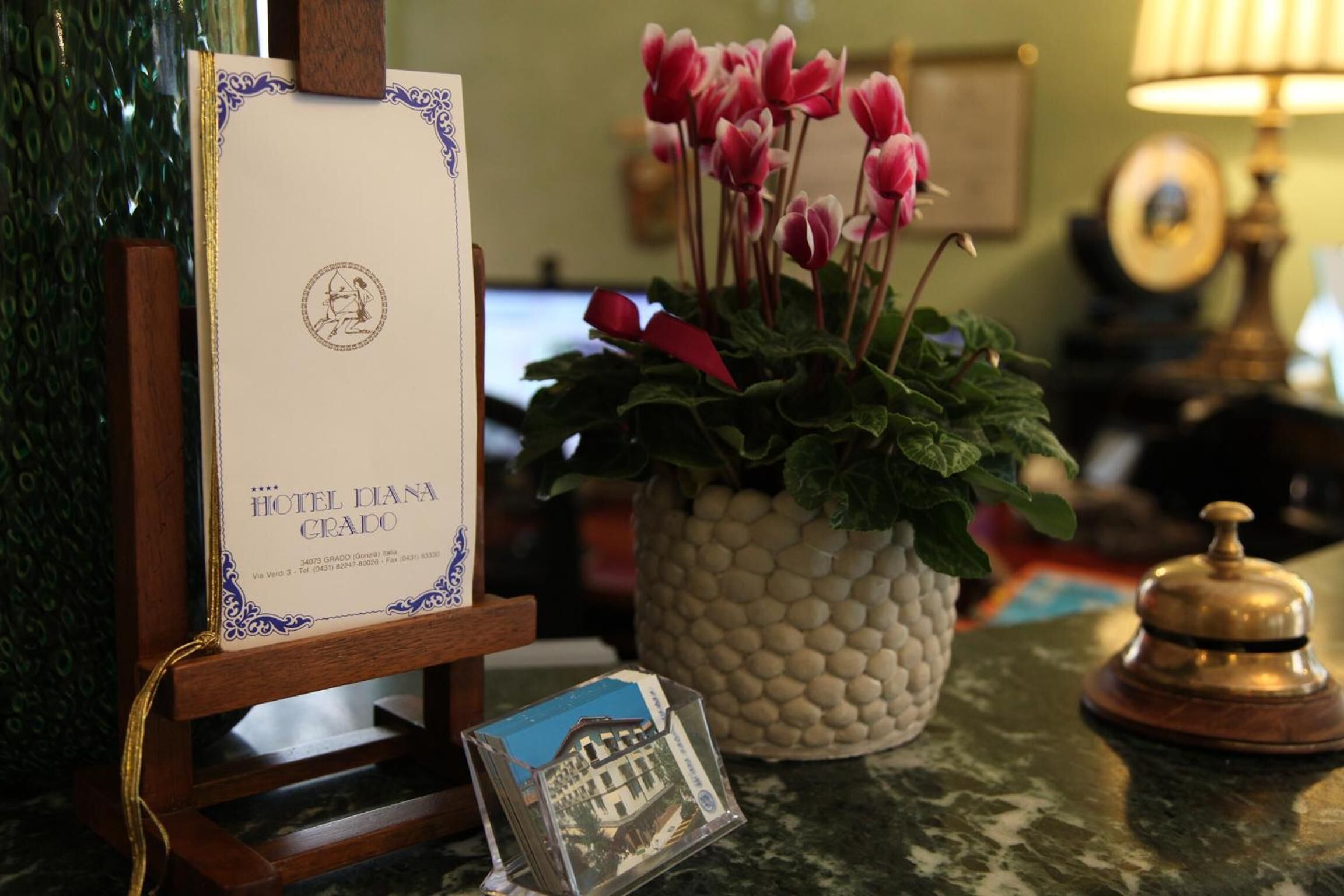 A hotel reception desk with a framed welcome sign, a potted plant with red flowers, a small framed picture, and a bell.