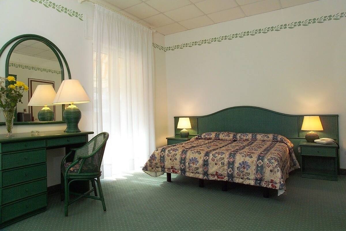 A bedroom with a green bed, two bedside lamps, a green desk with a chair, a mirror, and a window with white curtains.