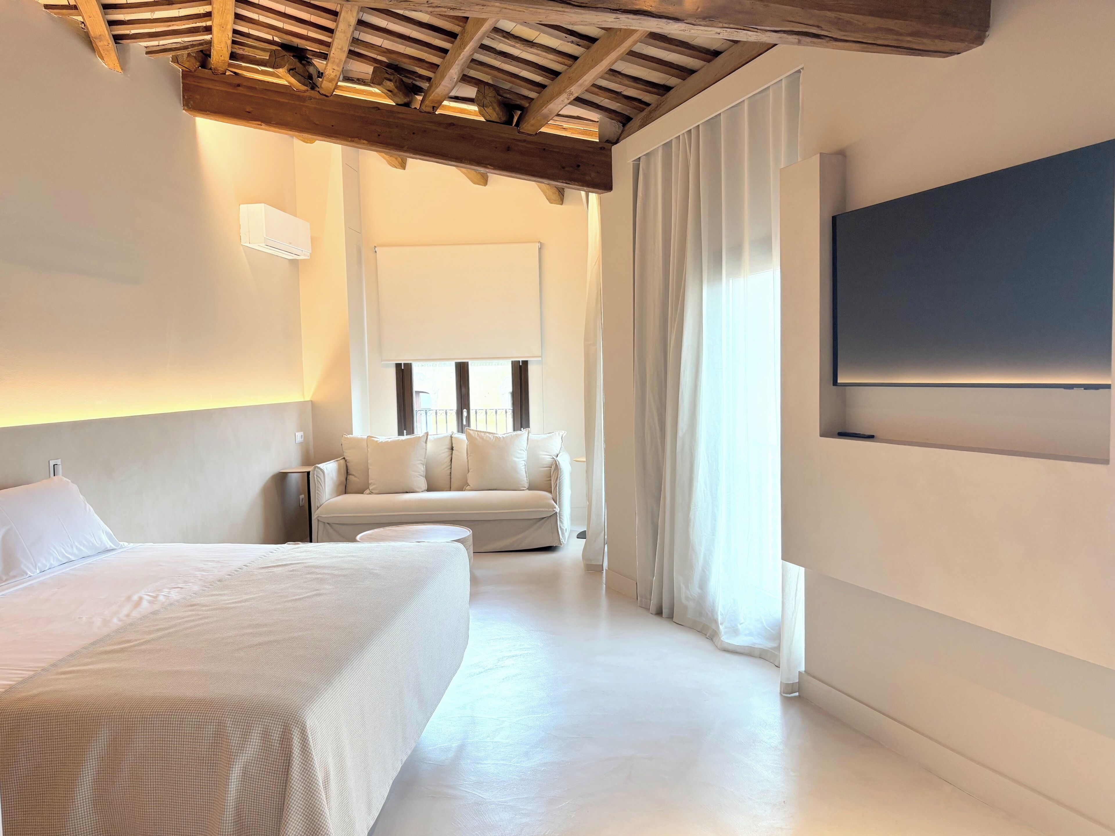 A modern hotel room with a large bed, a sofa, a TV mounted on the wall, and a ceiling with exposed wooden beams.