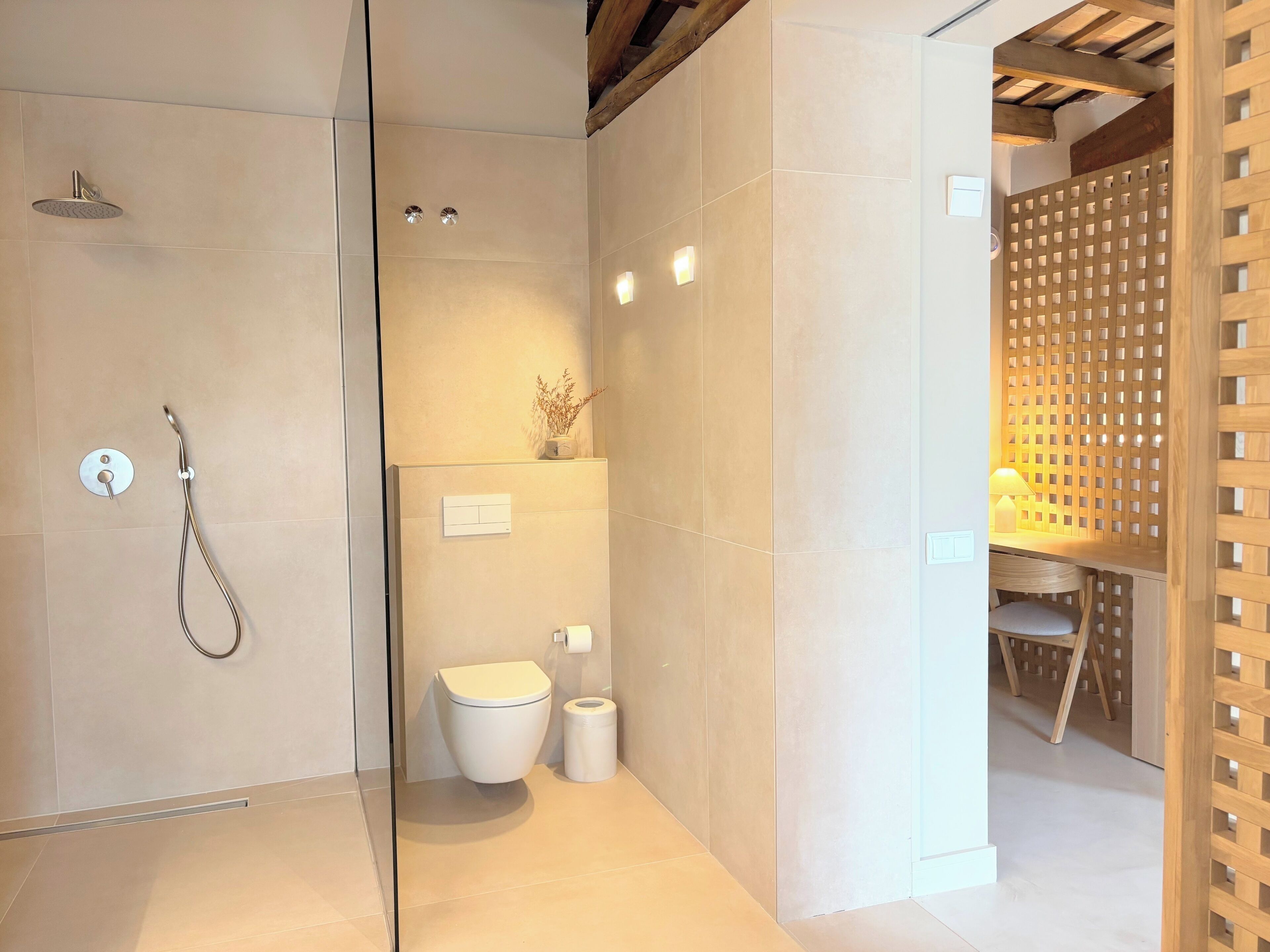 Un baño moderno con ducha de vidrio, inodoro y zona de lavamanos separada por un panel de celosía de madera.