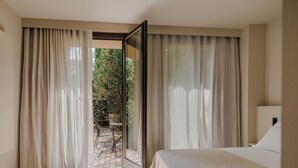 Een hotelkamer met een groot bed, uitzicht op een tuin door een glazen deur, en een klein tafeltje met stoelen buiten.
