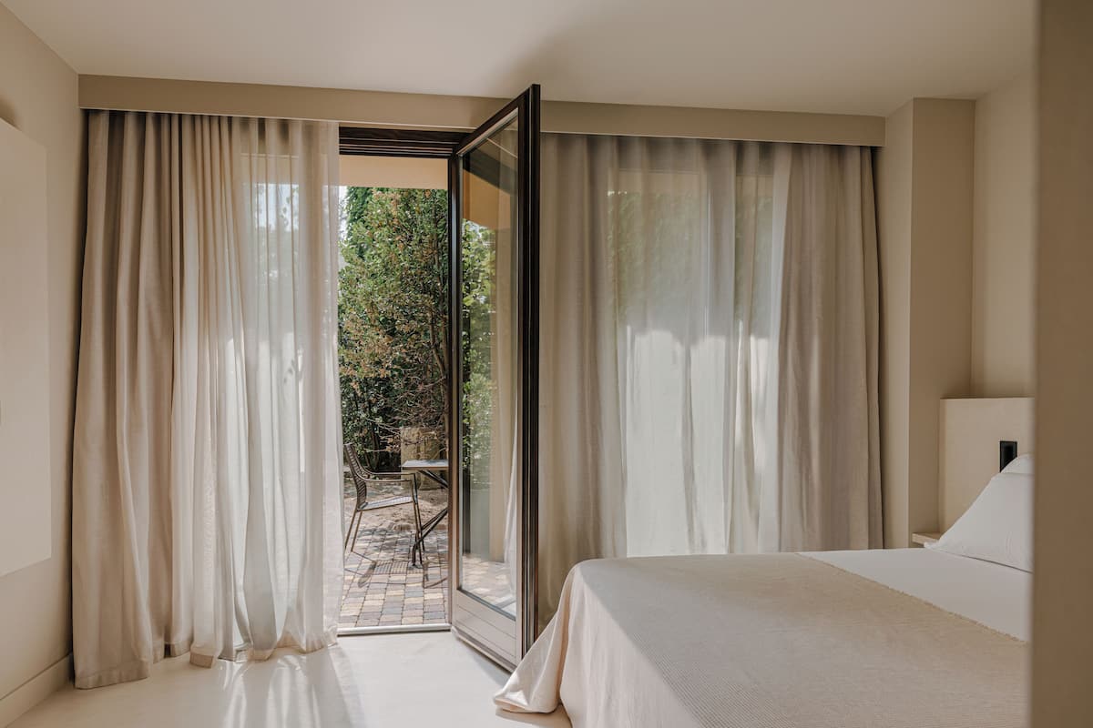 Une chambre d’hôtel avec un grand lit, une vue sur un jardin par une porte vitrée et une petite table avec des chaises à l’extérieur.