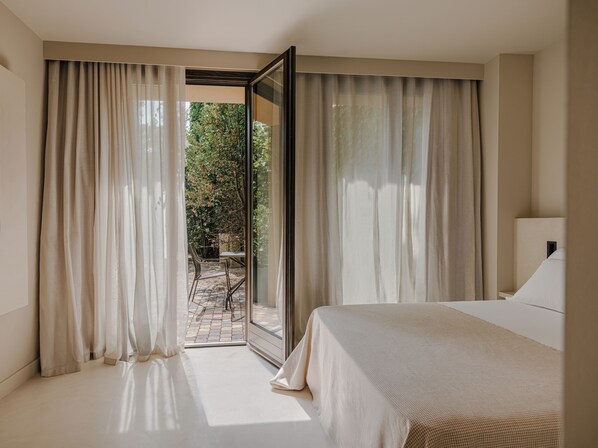 Habitación de hotel con una cama grande, vista de un jardín a través de un ventanal y una pequeña mesa con sillas afuera.