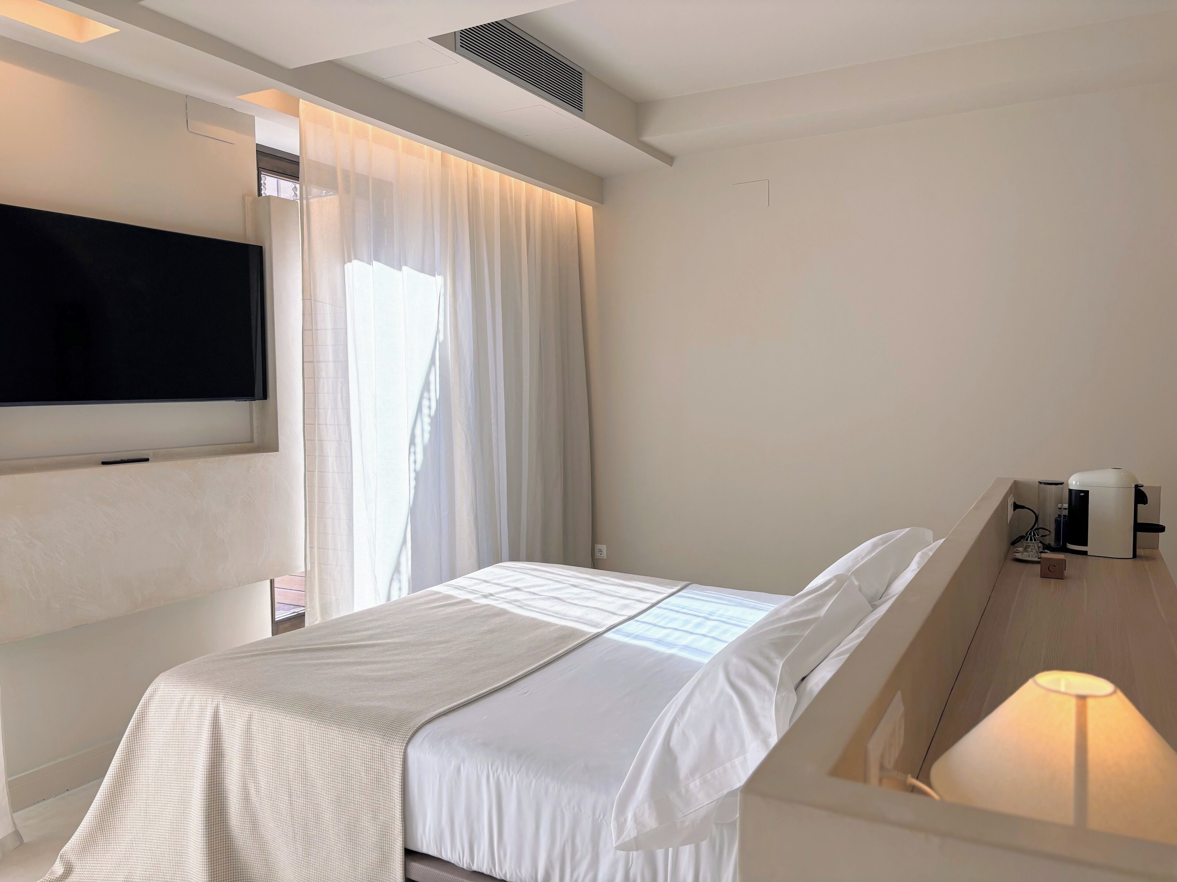 Una habitación de hotel moderna con una cama, un televisor de pantalla plana montado en la pared, una ventana con cortinas traslúcidas y una mesita de noche con lámpara.