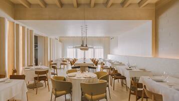 Un vaste espace dédié à la salle à manger, avec de nombreuses tables et chaises rondes, de hauts plafonds ornés de poutres en bois et de grandes fenêtres laissant entrer la lumière naturelle.