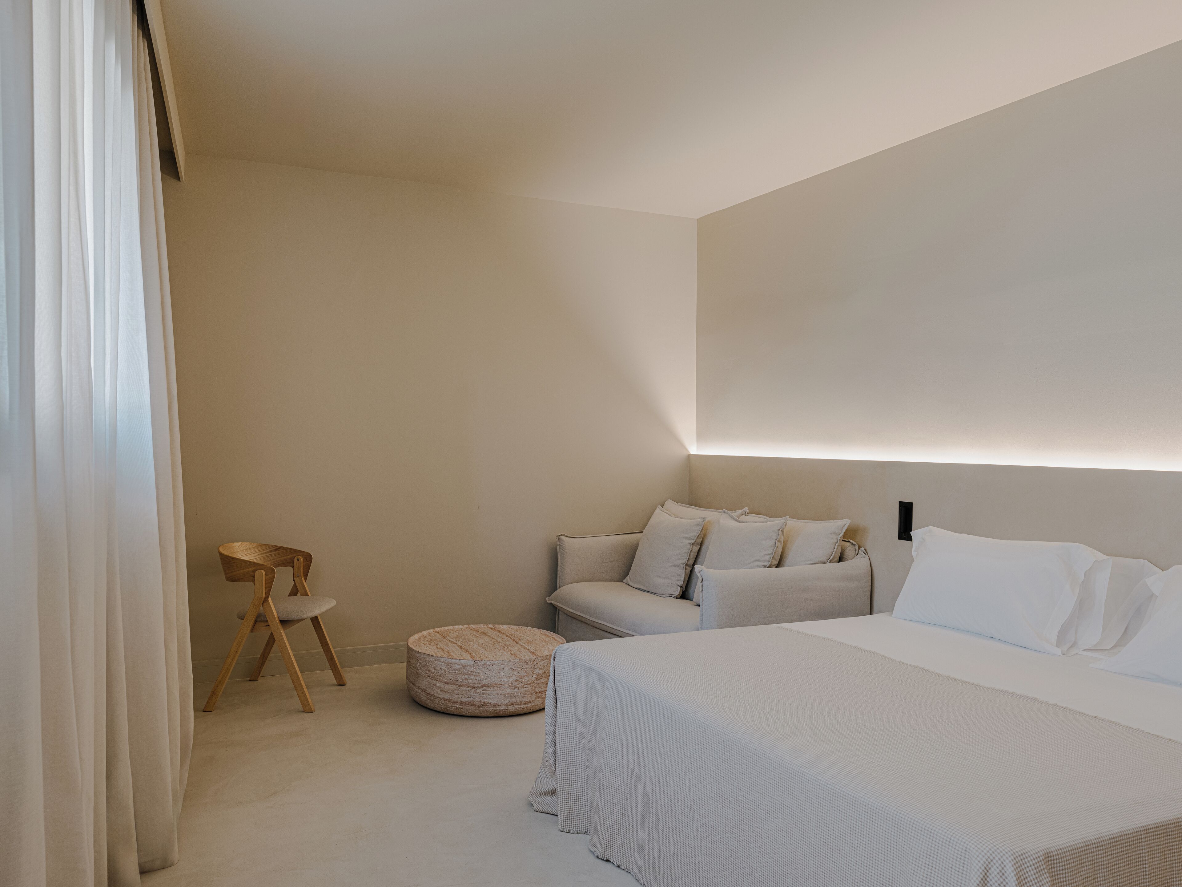 Un dormitorio minimalista con una cama, un sofá, un otomano redondo y una silla de madera.