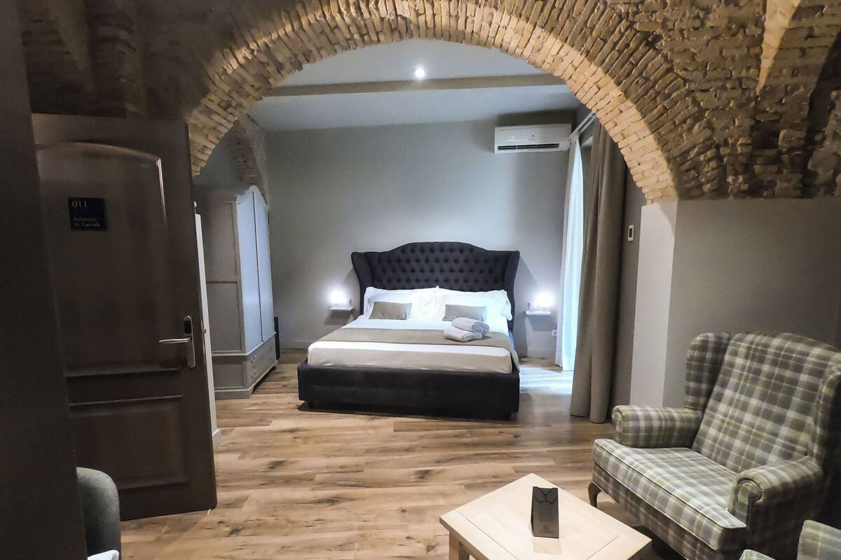 Une chambre d’hôtel avec un lit, un fauteuil à motifs à carreaux, une petite table et un plancher en bois.