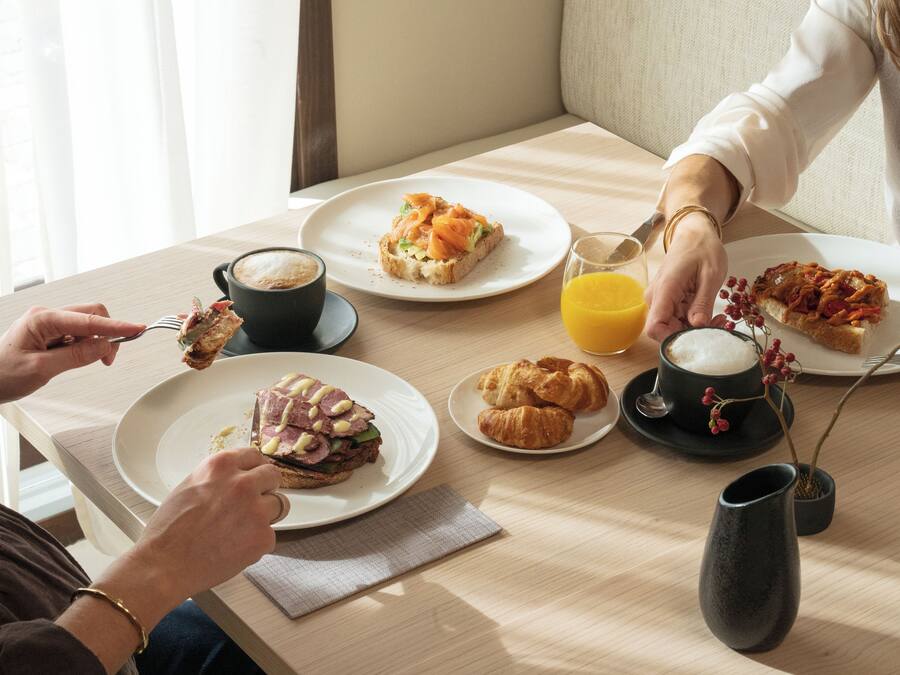 Aamiaispöytä, jossa on croissantteja, kinkkuvoileipä ja kuppi kahvia puupöydällä.