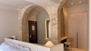 Une chambre d’hôtel avec une arche en briques, un lit avec du linge de maison blanc, un bureau avec une lampe et une armoire en bois.
