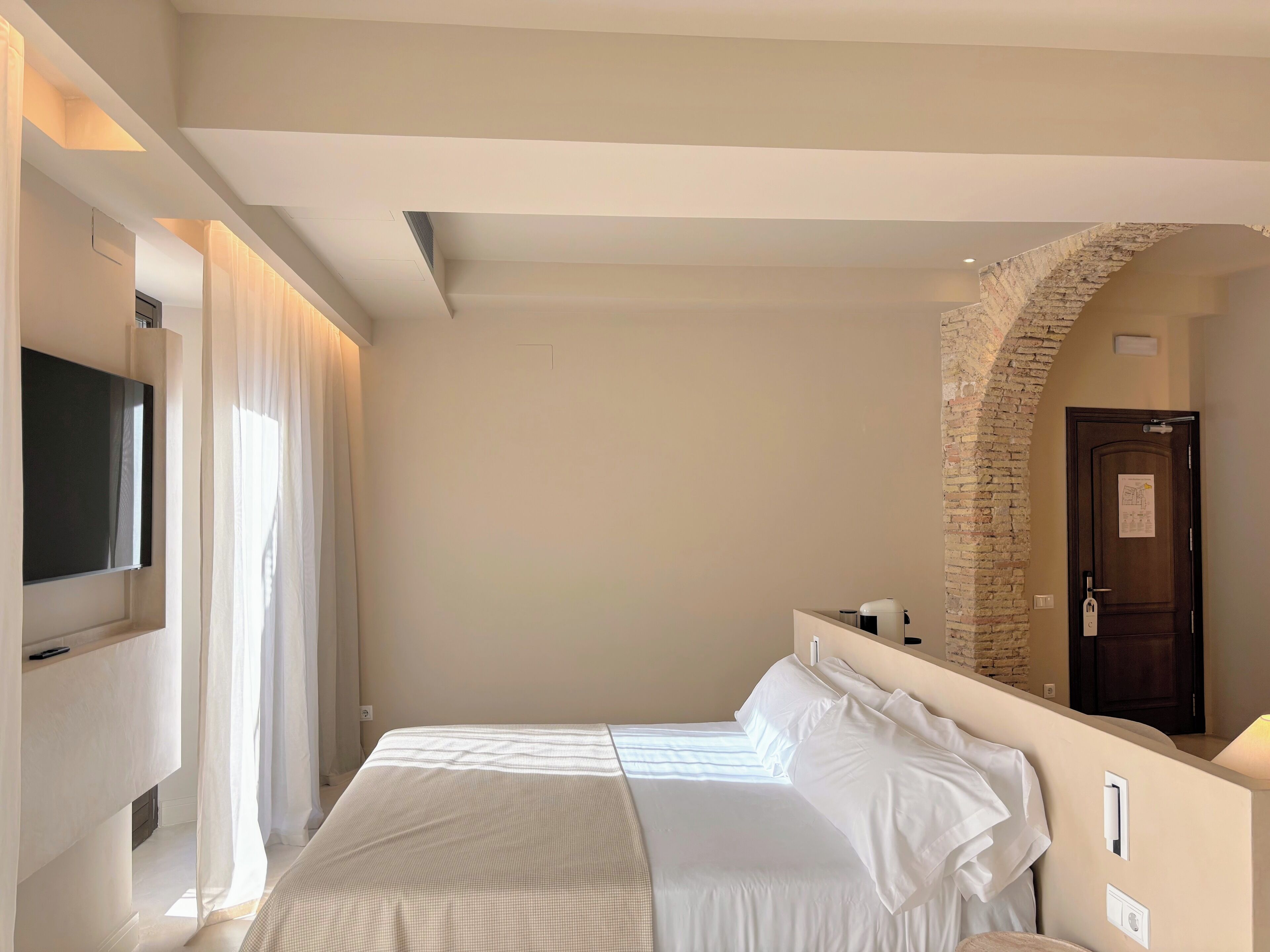 A hotel room with a bed, a TV mounted on the wall, a stone archway, and a door with a sign.