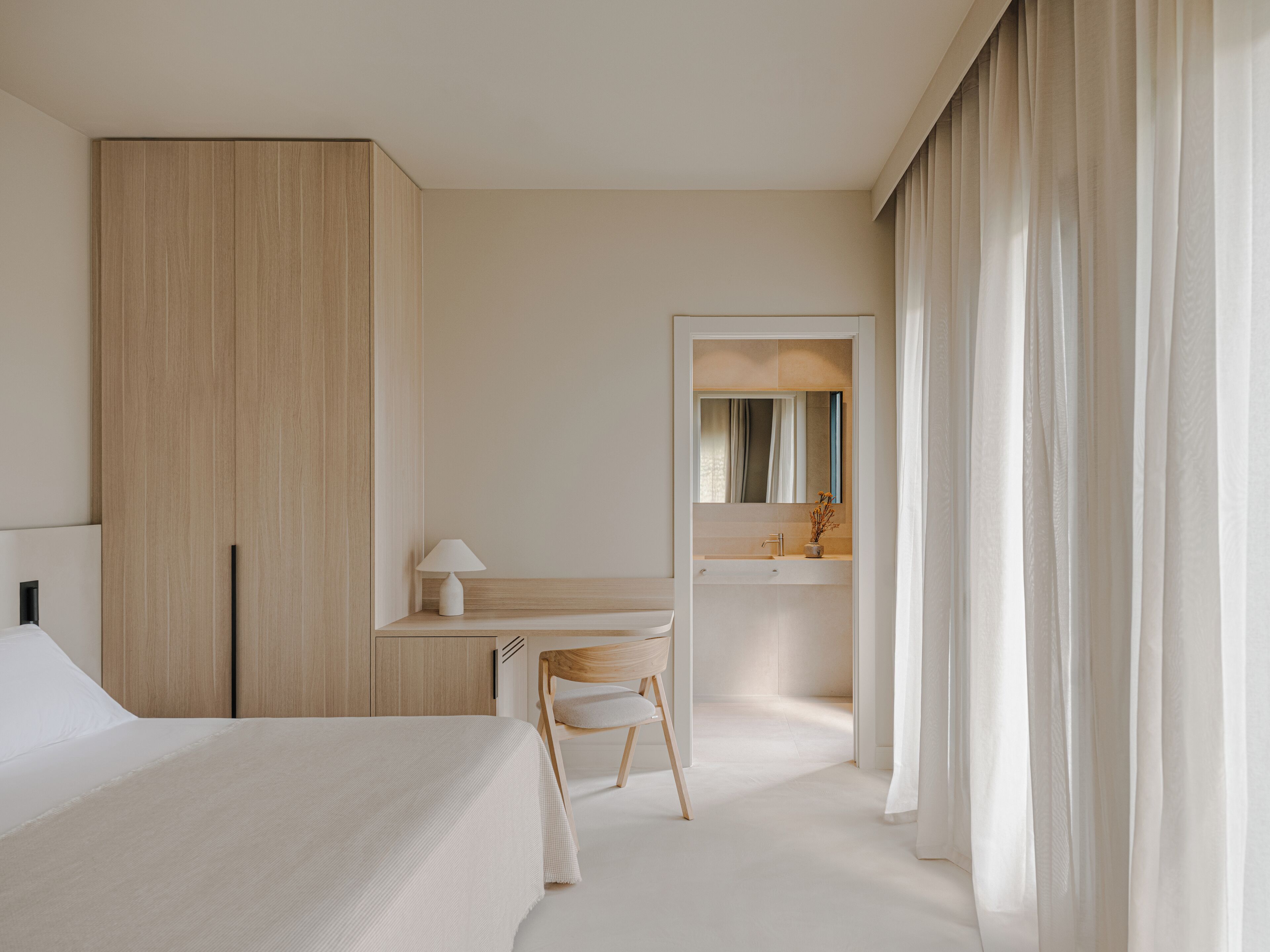 A modern bedroom with a large bed, a wooden wardrobe, a desk with a chair, and a bathroom visible through an open door.