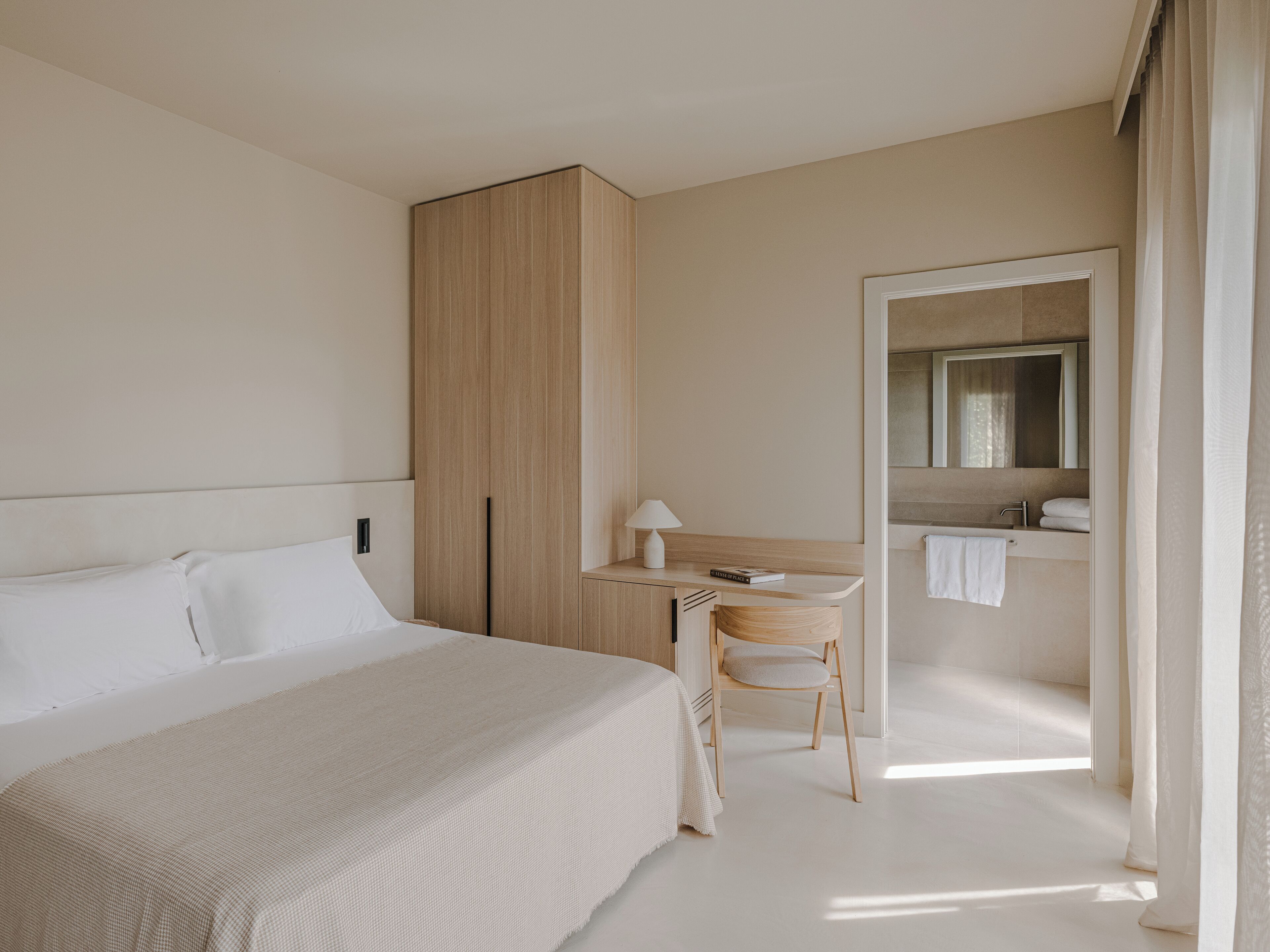 A hotel room with a bed, a desk, a chair, a wardrobe, and a bathroom with a mirror and towel rack.