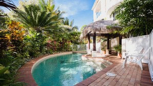 A swimming pool with a thatched roof gazebo, surrounded by lush tropical plants and a white fence. - Villa Beach Palm Cove (Clifton Beach)