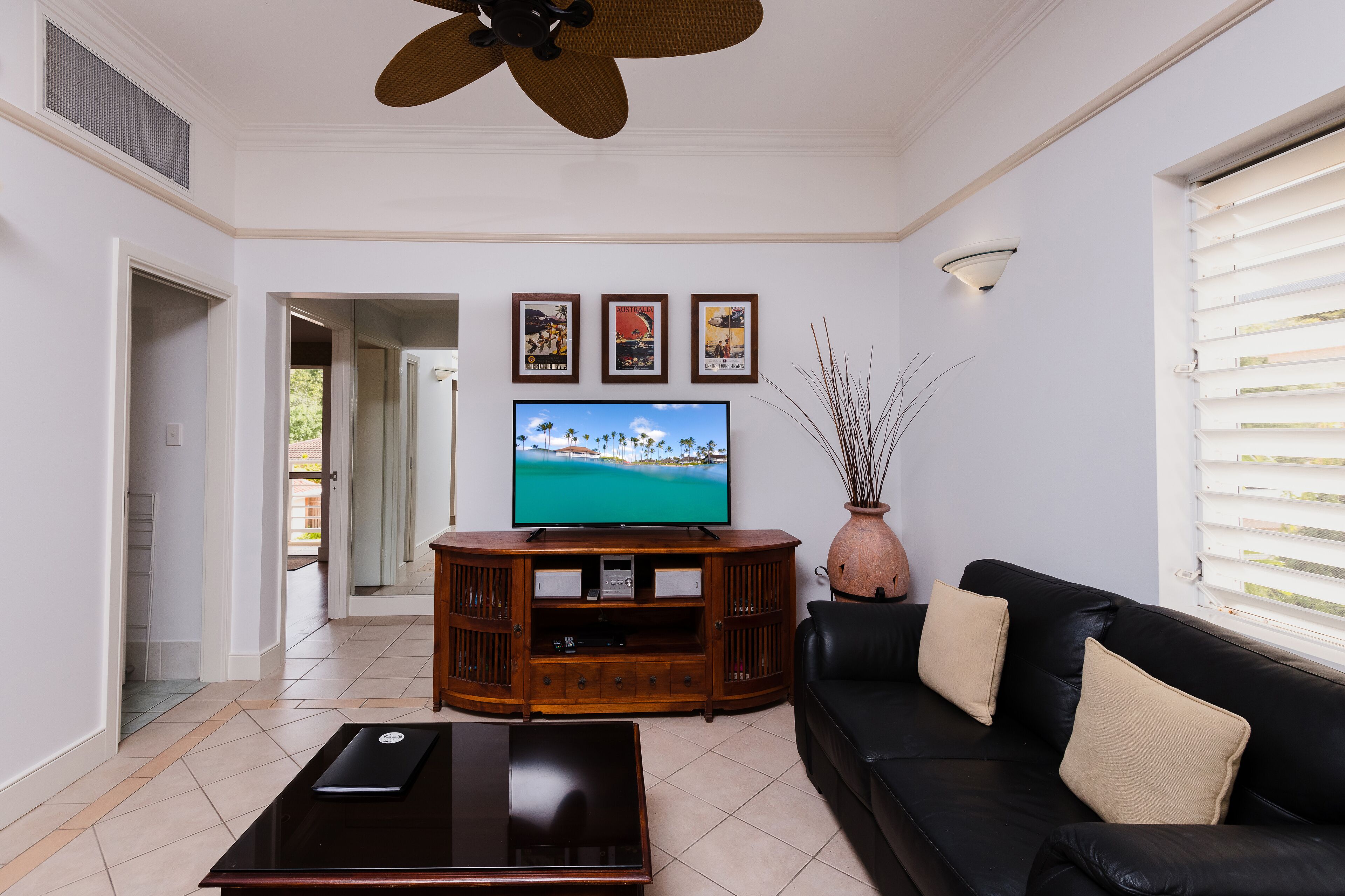 A living room with a television, a wooden entertainment unit, a black leather sofa, and a vase with branches.