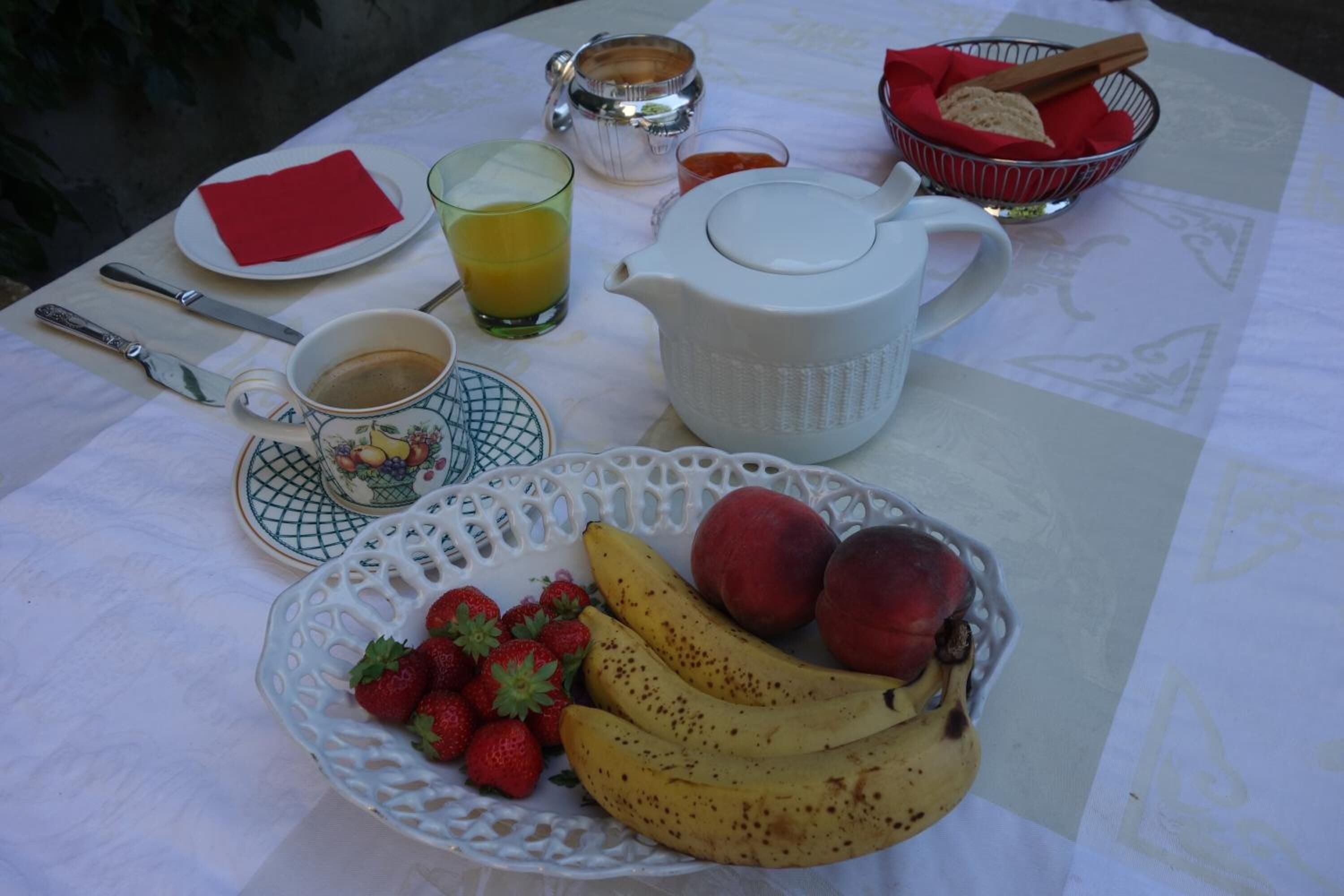 Een ontbijttafel met een fruitschaal, een koffiekop, een theepot en een glas sinaasappelsap.