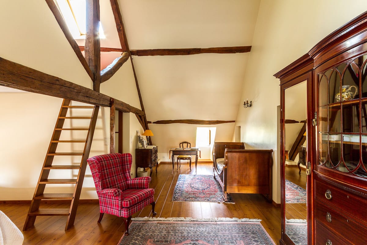 Ein Schlafzimmer im Loft-Stil mit freiliegenden Holzbalken, einer Leitern und einem roten karierten Sessel, einer Holzkommode und einem großen Spiegel.