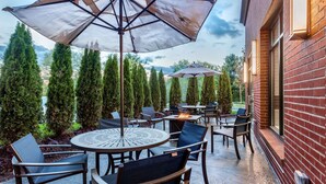 An outdoor patio area with round tables and chairs, umbrellas, and a brick building in the background. - Hampton Inn & Suites Kittery-Portsmouth (Kittery)