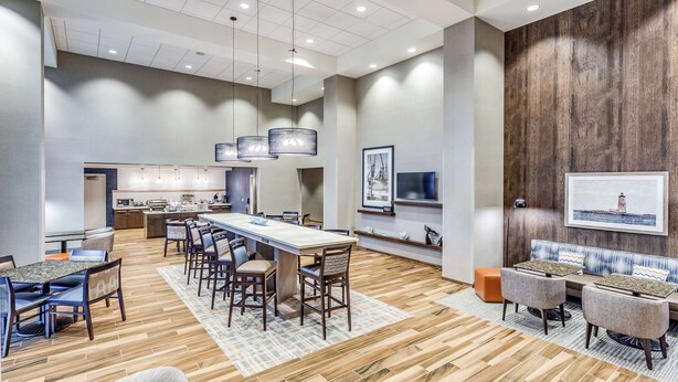 A modern hotel lobby with a central dining area, a kitchen, and a seating area with a wooden accent wall and artwork. - Hampton Inn & Suites Kittery-Portsmouth (Kittery)