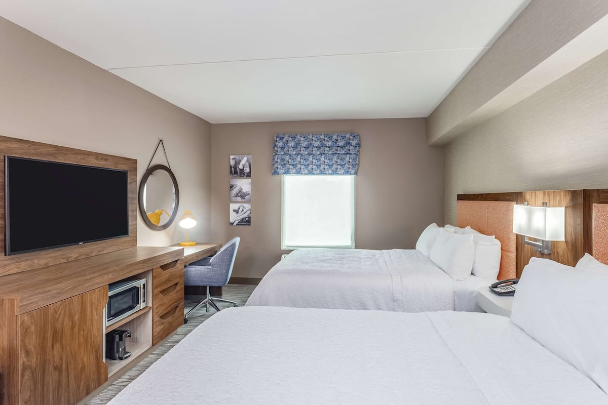 Quarto de hotel com duas camas, televisão, secretária com candeeiro, cadeira e janela com vista.