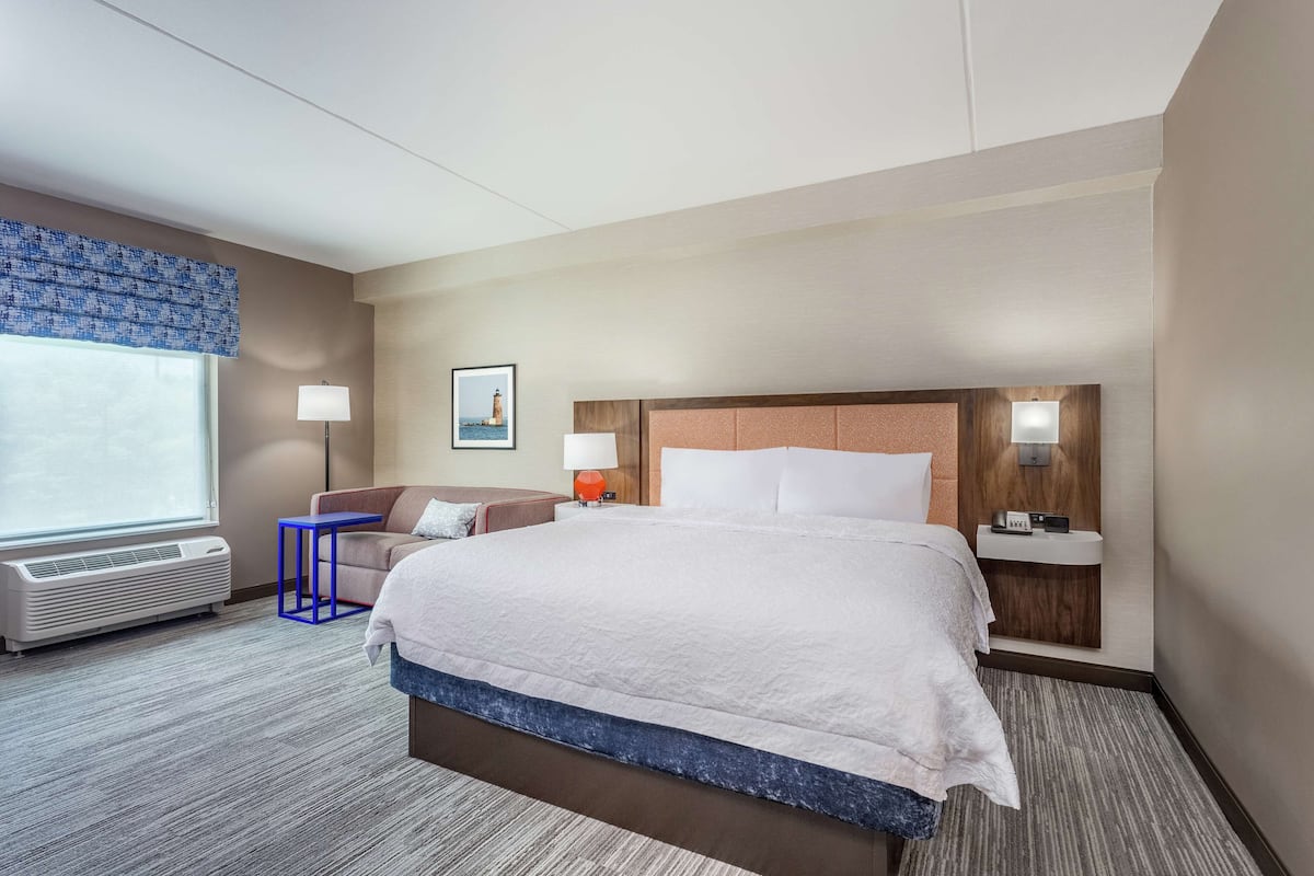 Quarto de hotel com uma cama grande, uma escrivaninha, uma cadeira, um abajur e um quadro na parede.