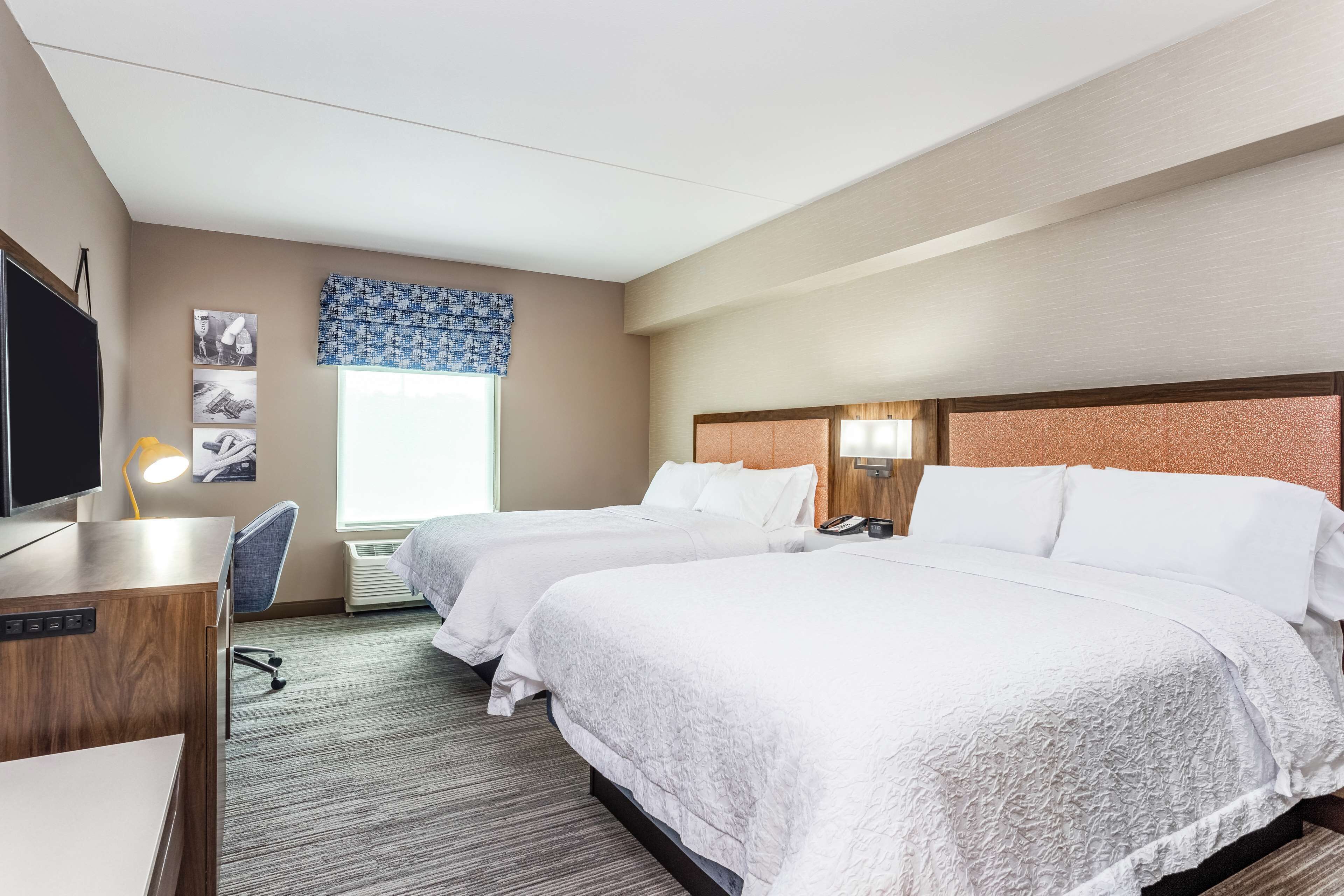 A hotel room with two beds, a desk with a computer, a TV, and a window with a blue curtain.